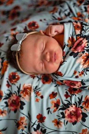 A sleeping newborn wearing a white bow headband rests on floral patterned fabric.