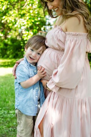 A young child embraces a pregnant woman in a pink dress outdoors on a sunny day.