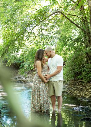 A romantic couple embraces while standing in a creek surrounded by lush green foliage.