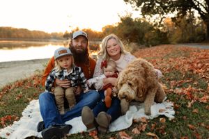 Family and their fluffy dog sit together on a blanket by the lake during fall.