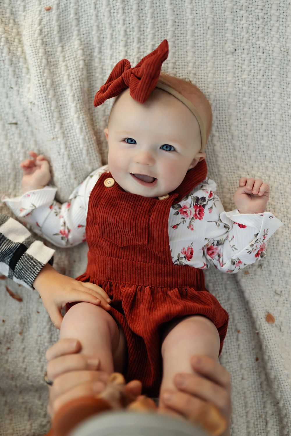Baby wearing red corduroy outfit with matching bow smiles while lying on textured blanket.