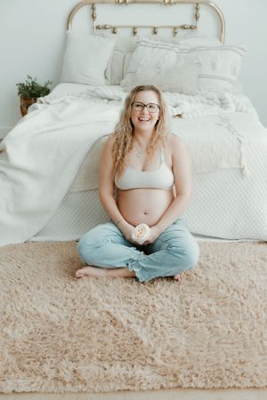 A pregnant person in casual attire sits cross-legged on a plush carpet in front of a white bed with decorative pillows.