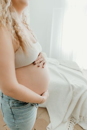 Close-up view of pregnant belly with hands cradling the bump, wearing jeans and white top in soft natural light.