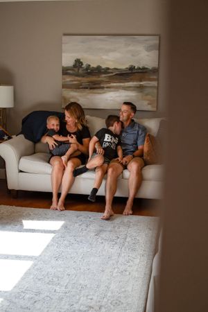 A family shares a tender moment together on a white couch beneath landscape artwork in a sunlit living room.
