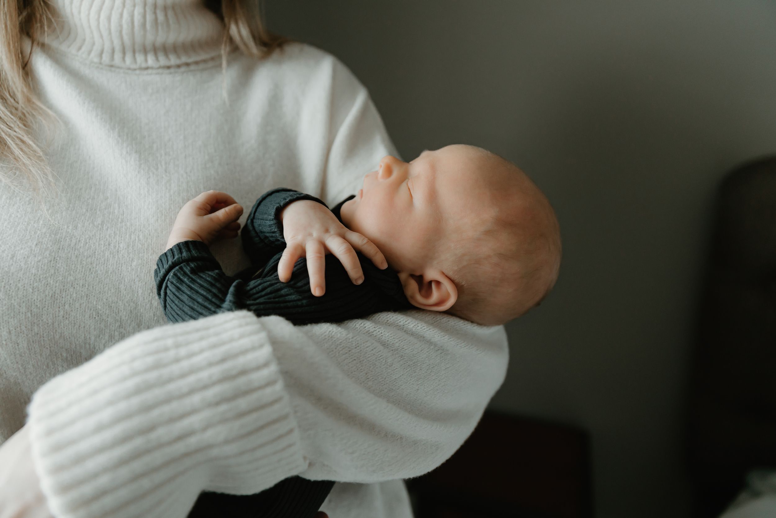A sleeping baby in dark clothing rests peacefully against someone wearing a cozy white turtleneck sweater.