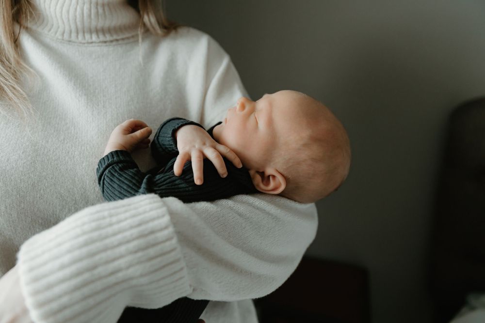A sleeping baby in dark clothing rests peacefully against someone wearing a cozy white turtleneck sweater.