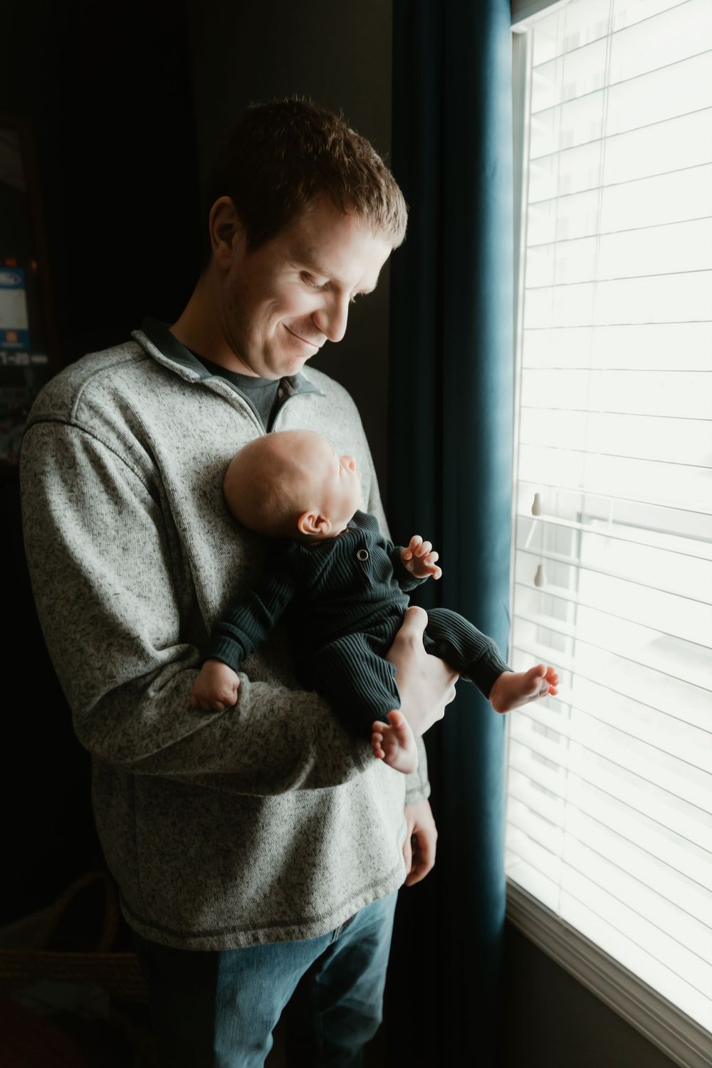 Parent in gray sweatshirt holding infant baby by window with natural lighting casting soft shadows.