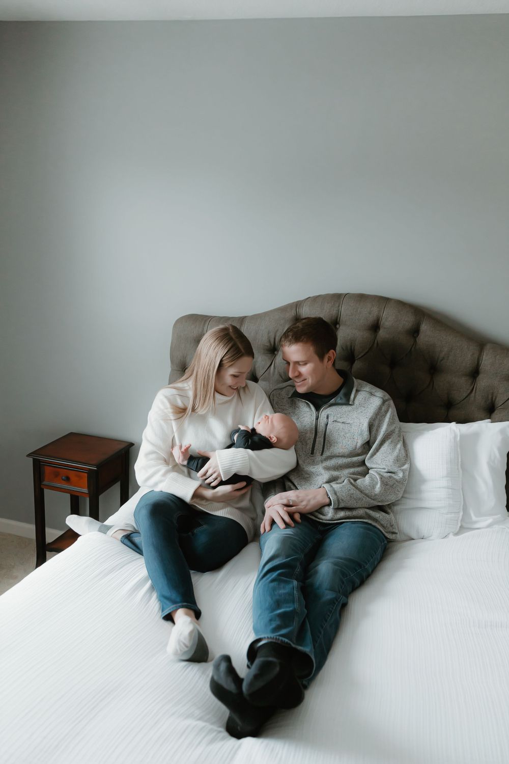 A couple sits together on a bed sharing an intimate moment with their newborn baby in a cozy bedroom setting.