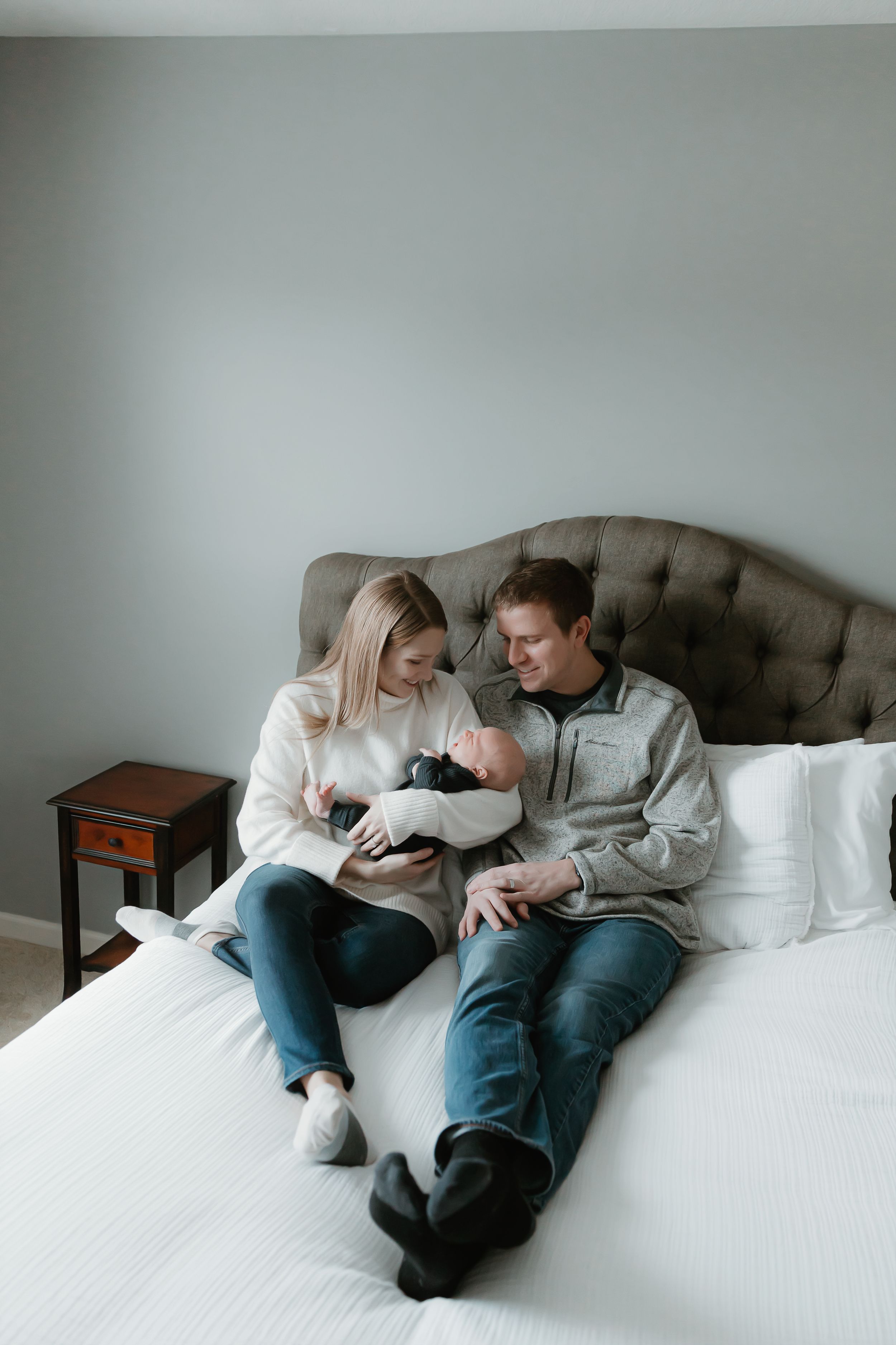 A couple sits together on a bed sharing an intimate moment with their newborn baby in a cozy bedroom setting.