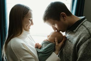 A couple shares a tender moment with their newborn baby near a window in soft natural light.