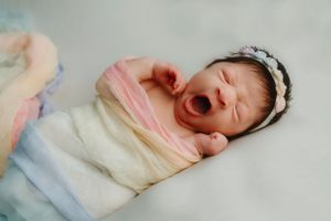 Sleepy newborn baby wrapped in soft pastel blankets yawns while lying on white bedding.