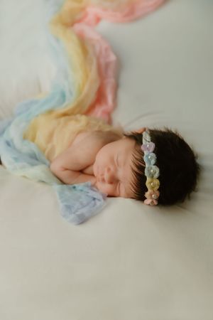 A sleeping newborn wearing a delicate floral headband rests on pastel rainbow-colored fabric.