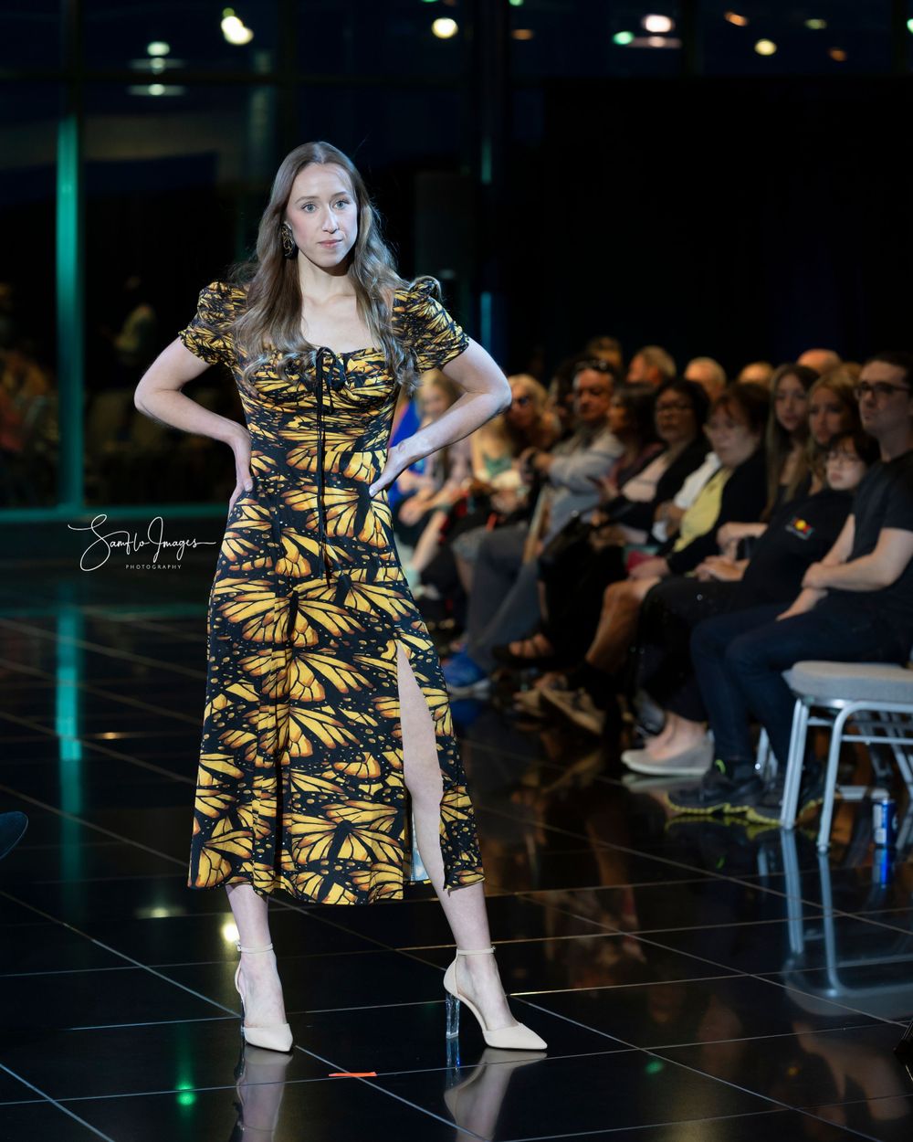 Model walks runway in black and gold butterfly print dress with side slit and white heels during fashion show.