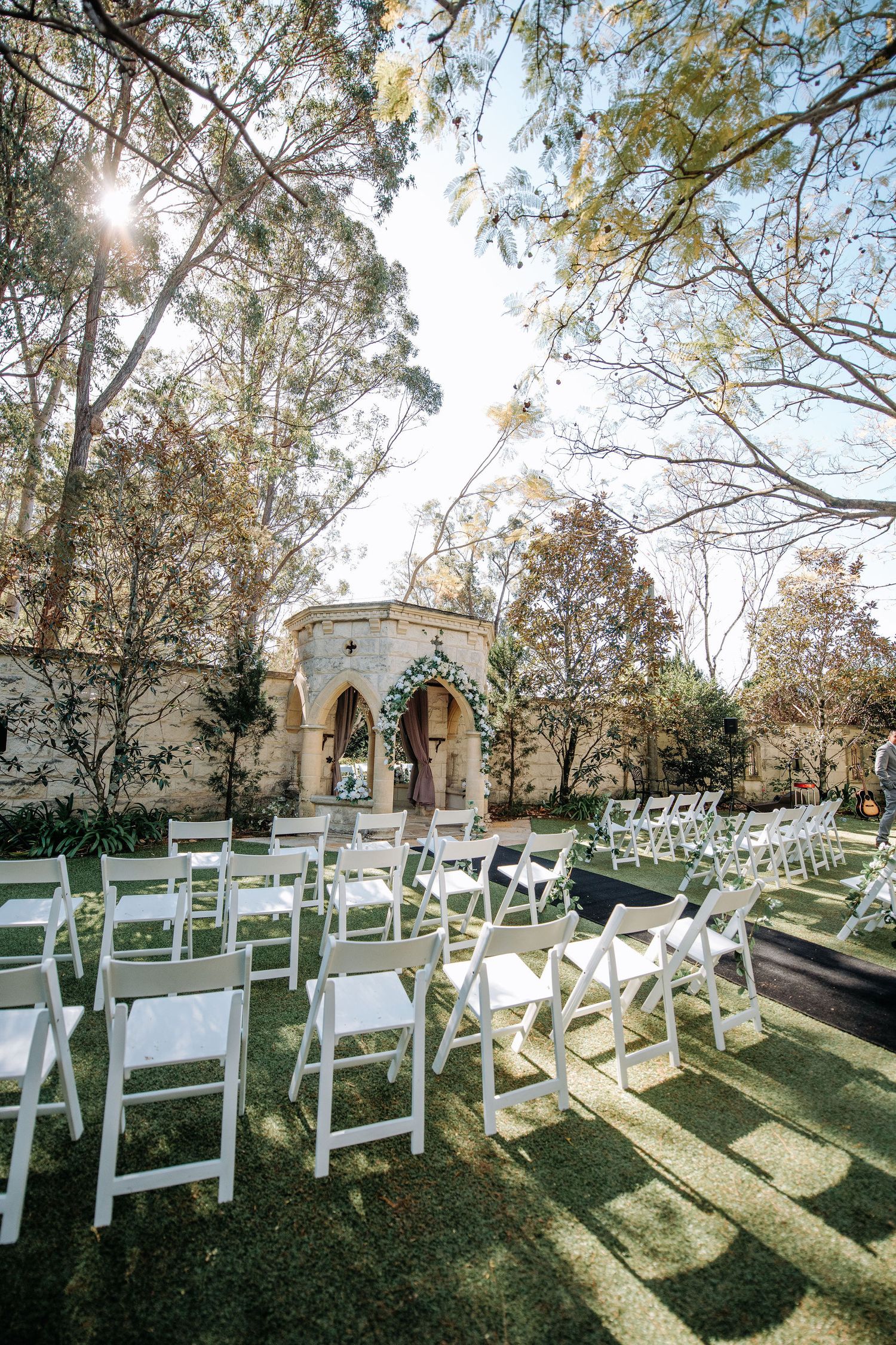 Outdoor wedding ceremony setup with white chairs under sunlit trees.