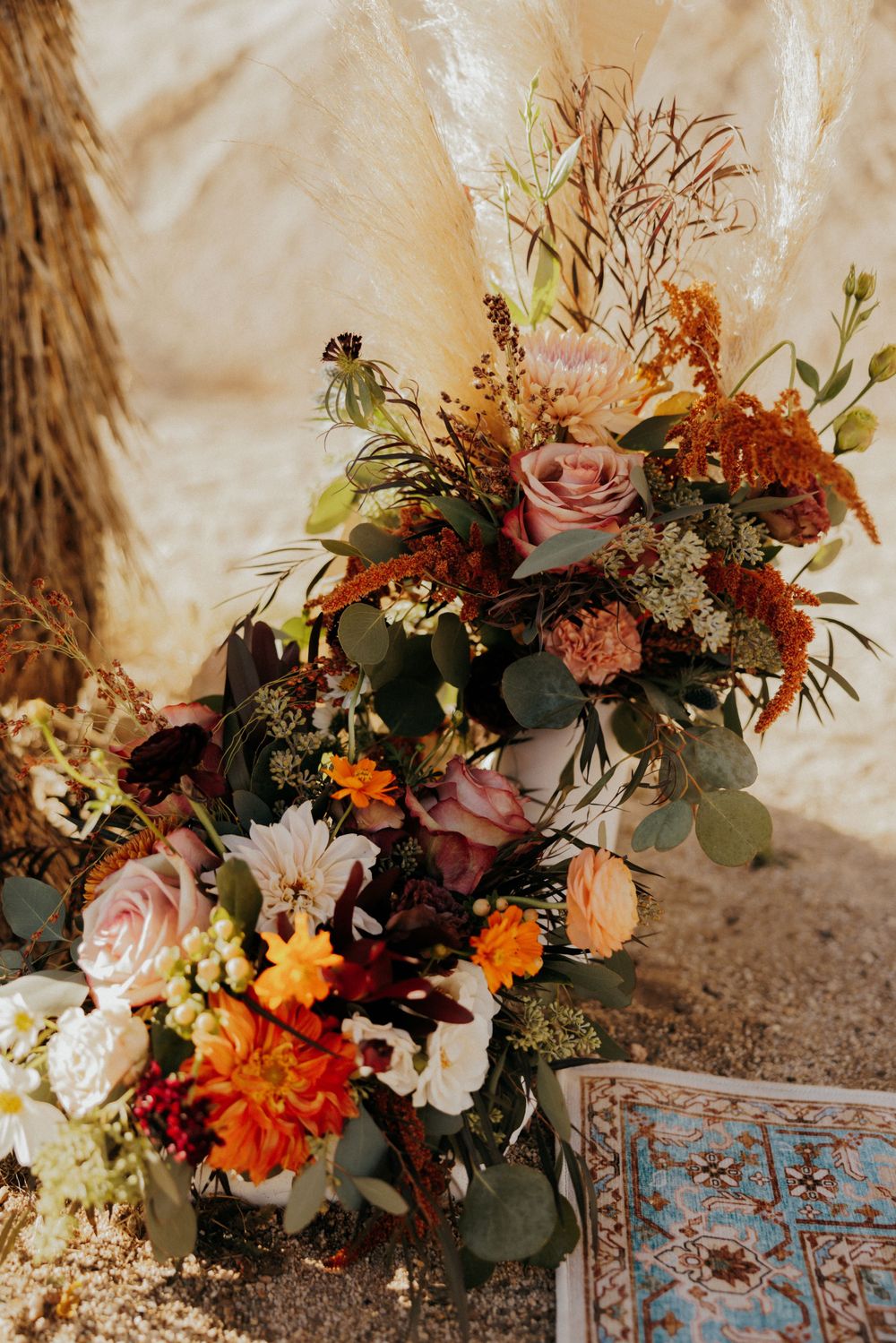 Romantic autumn wedding floral arrangement with pink roses, orange dahlias, and eucalyptus leaves against a beige backdrop.