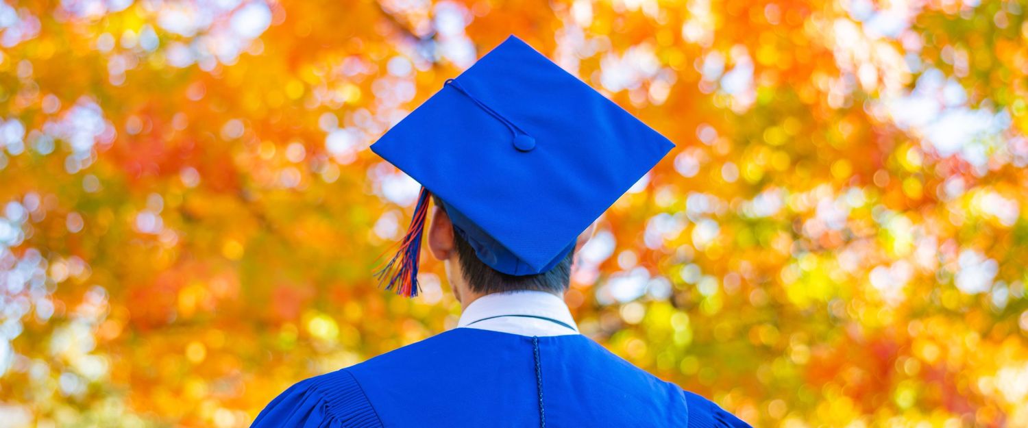 University of Illinois graduating student with a cap and gown looking up at fall leaves