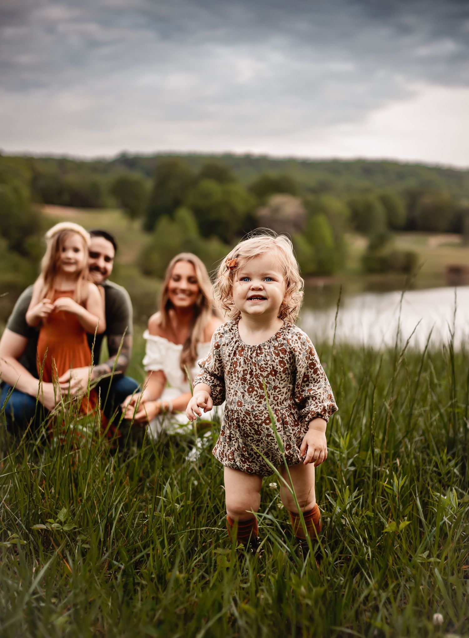 A small child wearing a floral dress runs joyfully through tall grass in a field on a cloudy day.