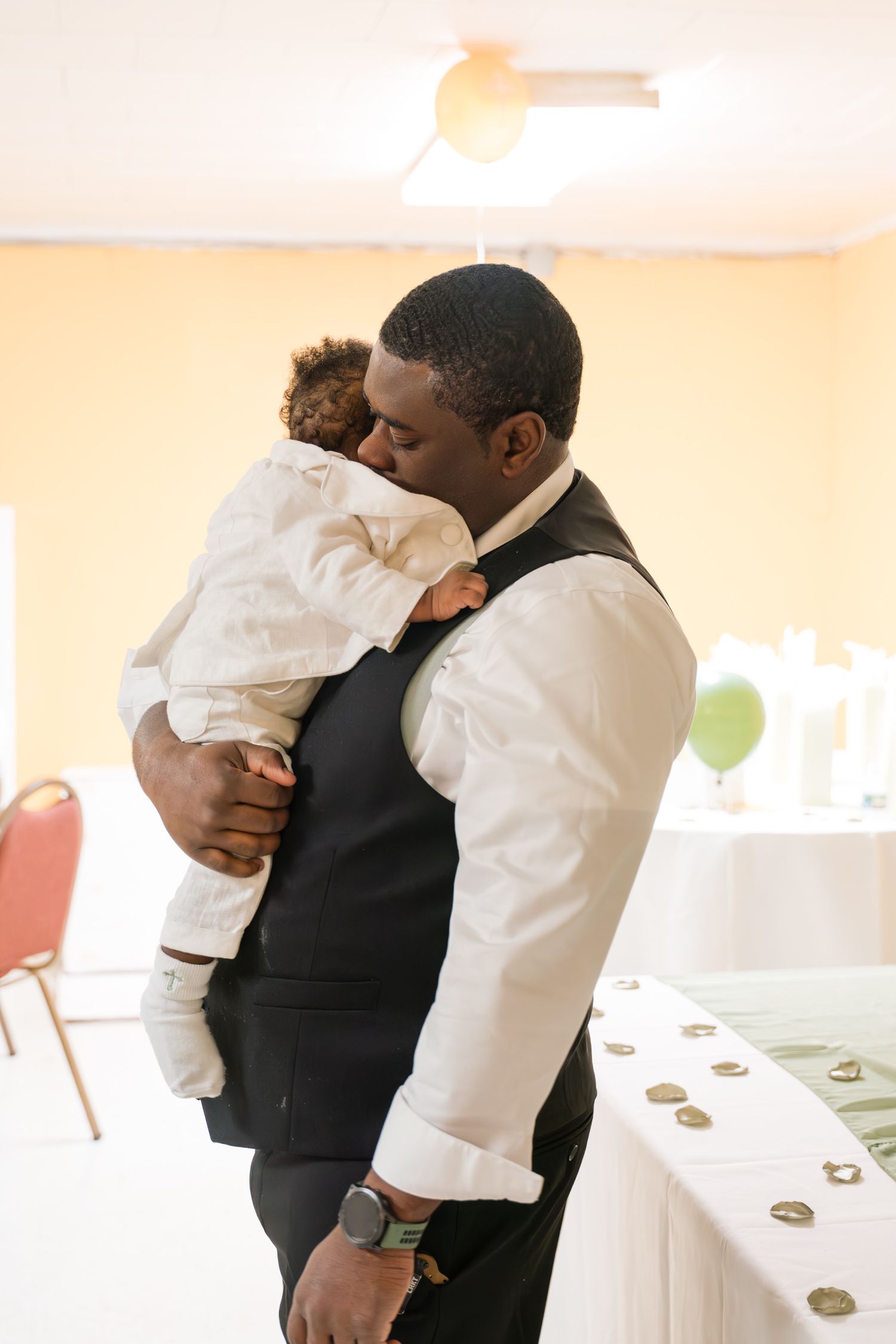 Elegantly dressed person in vest and white shirt holds small child in formal white outfit.