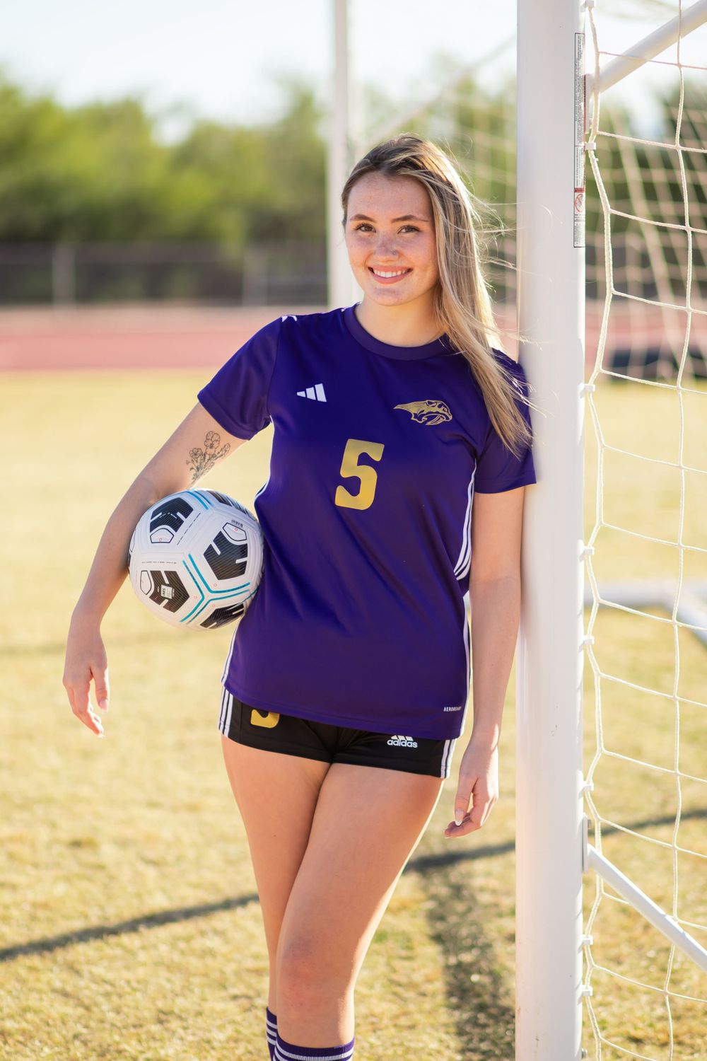 Soccer player in purple jersey poses by goal post on sunny field.