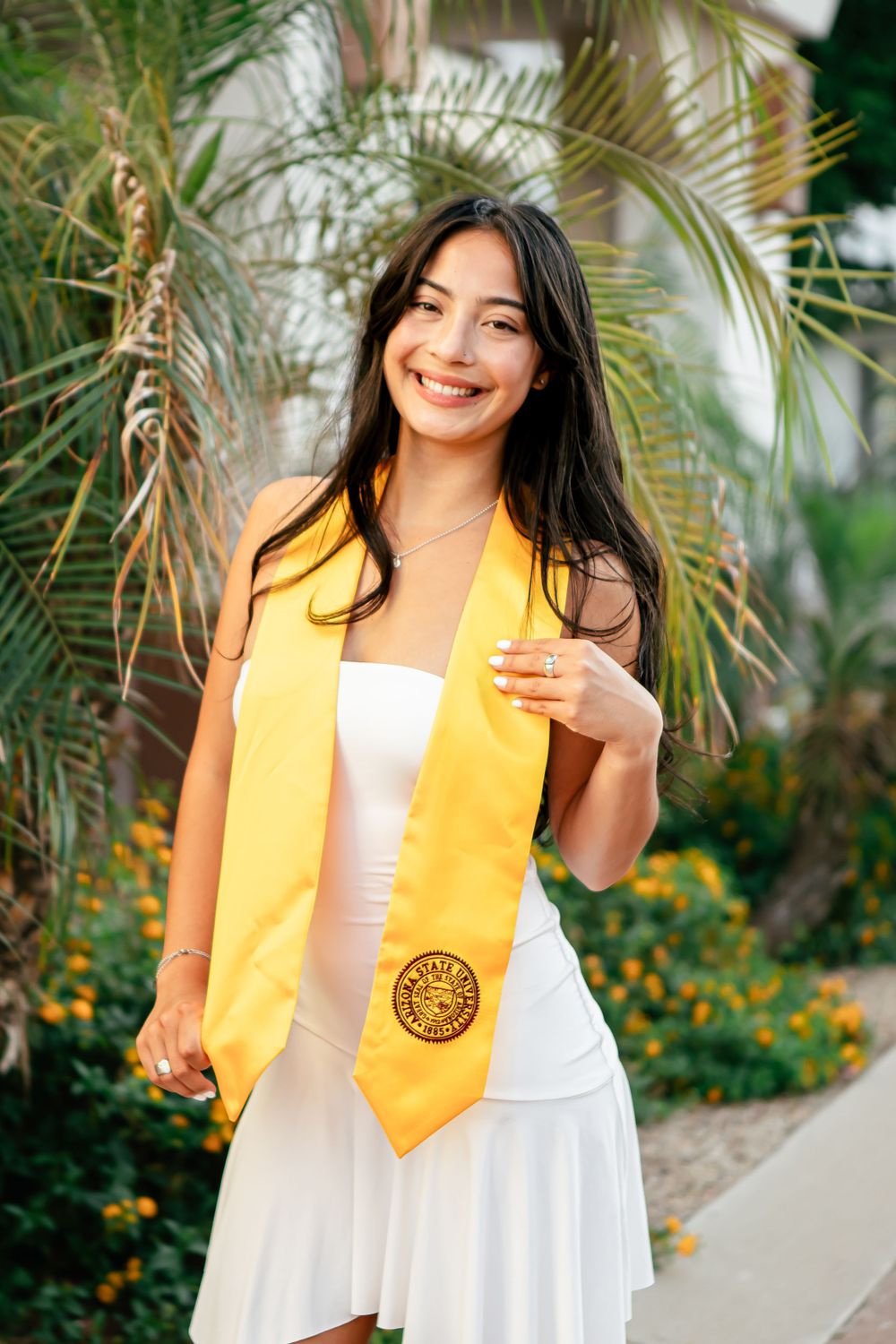 Graduate in yellow stole and white dress poses among tropical palm trees.