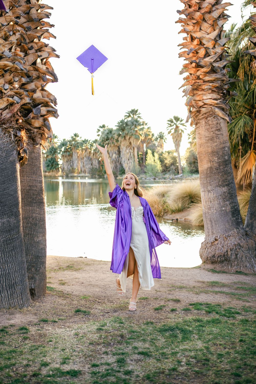 Woman in purple robe twirls by palm trees near a lake at sunset.