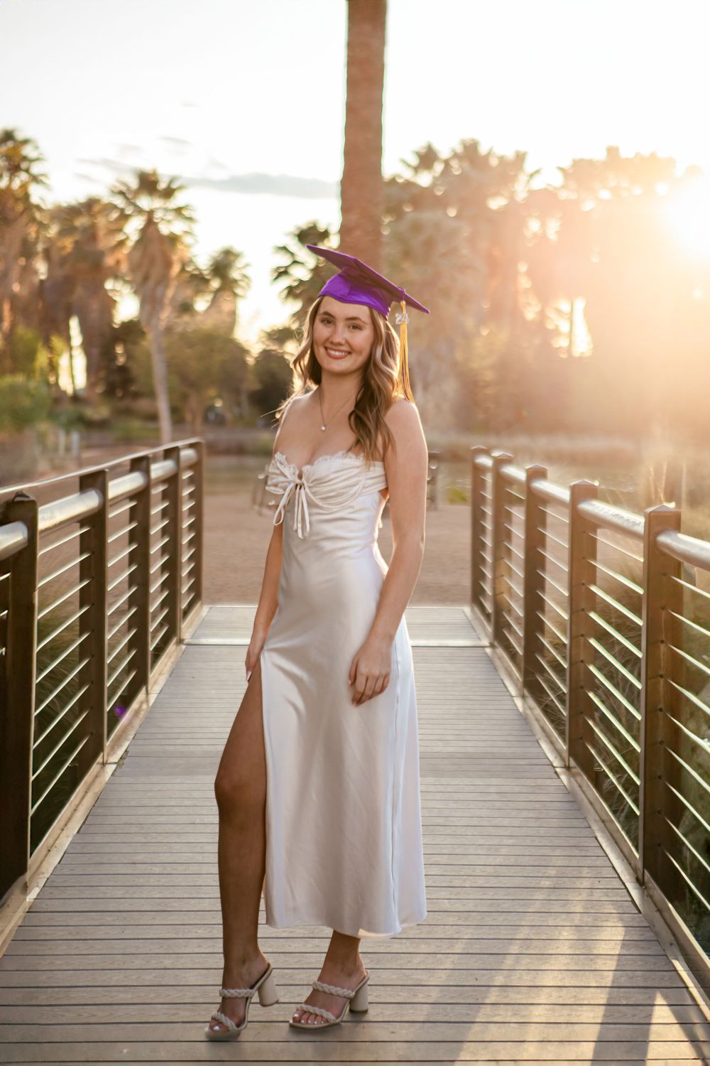 Woman in white dress and purple cap poses on wooden boardwalk at sunset.