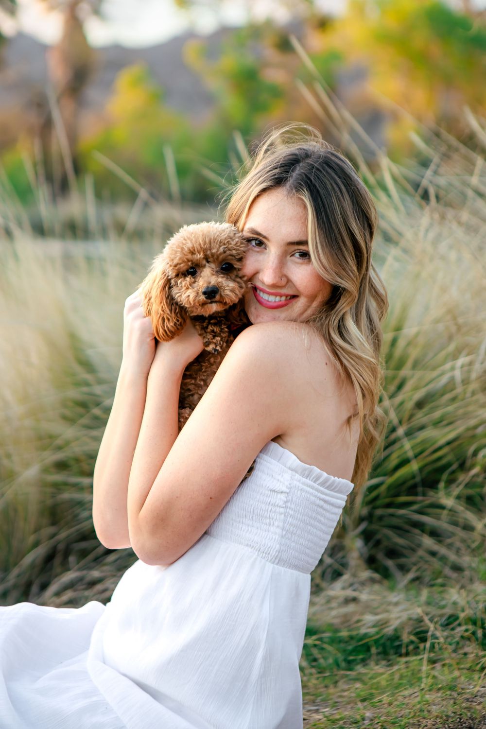 Woman in white dress cuddles small brown poodle outdoors.