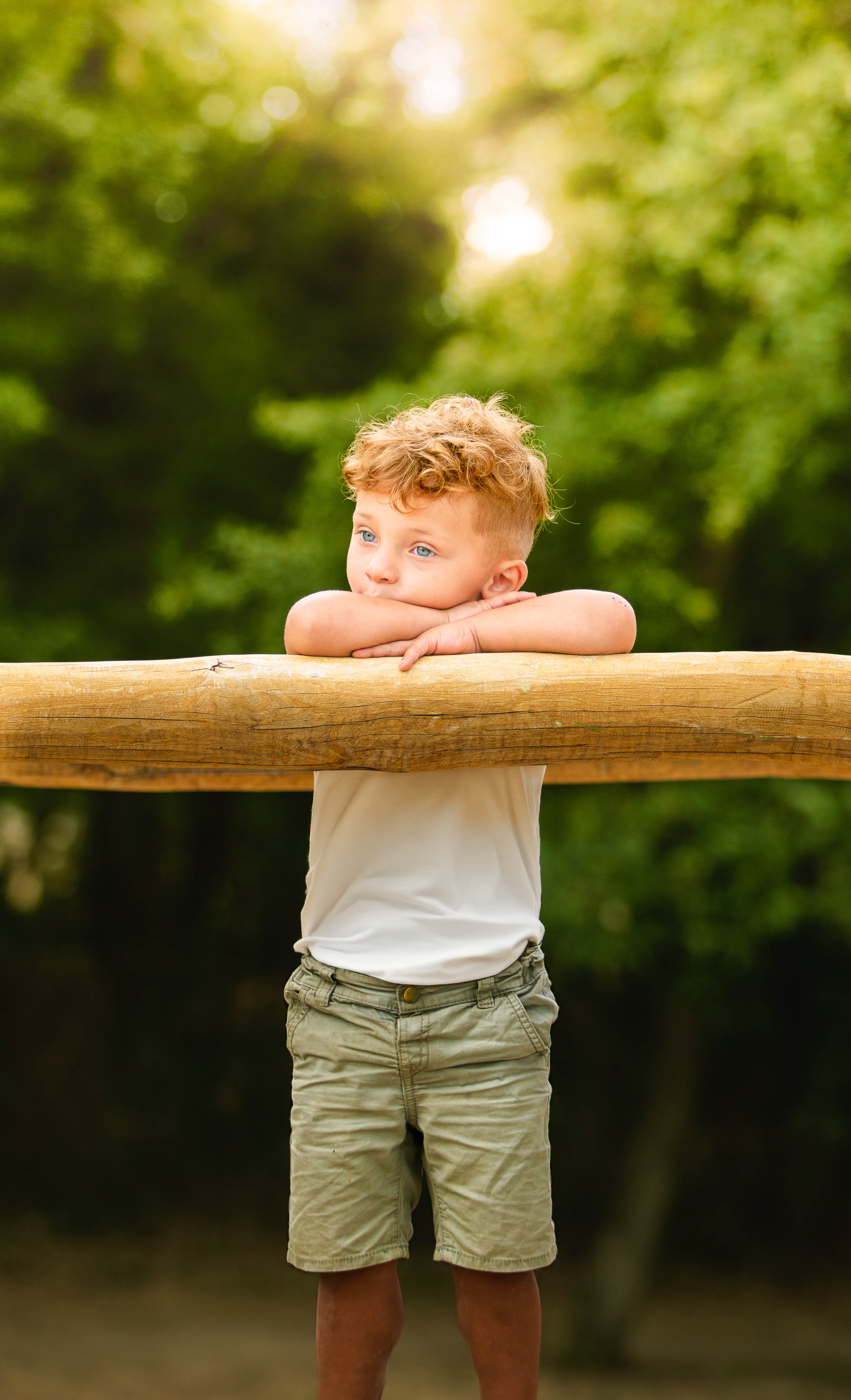 A young child in a white shirt and khaki shorts leans thoughtfully on a wooden railing in a sunlit garden setting.