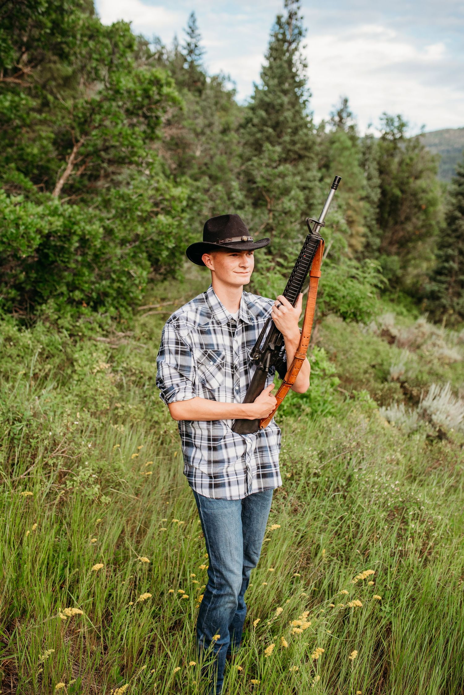 Person in plaid shirt and cowboy hat holding a rifle stands in tall grass with mountains behind.