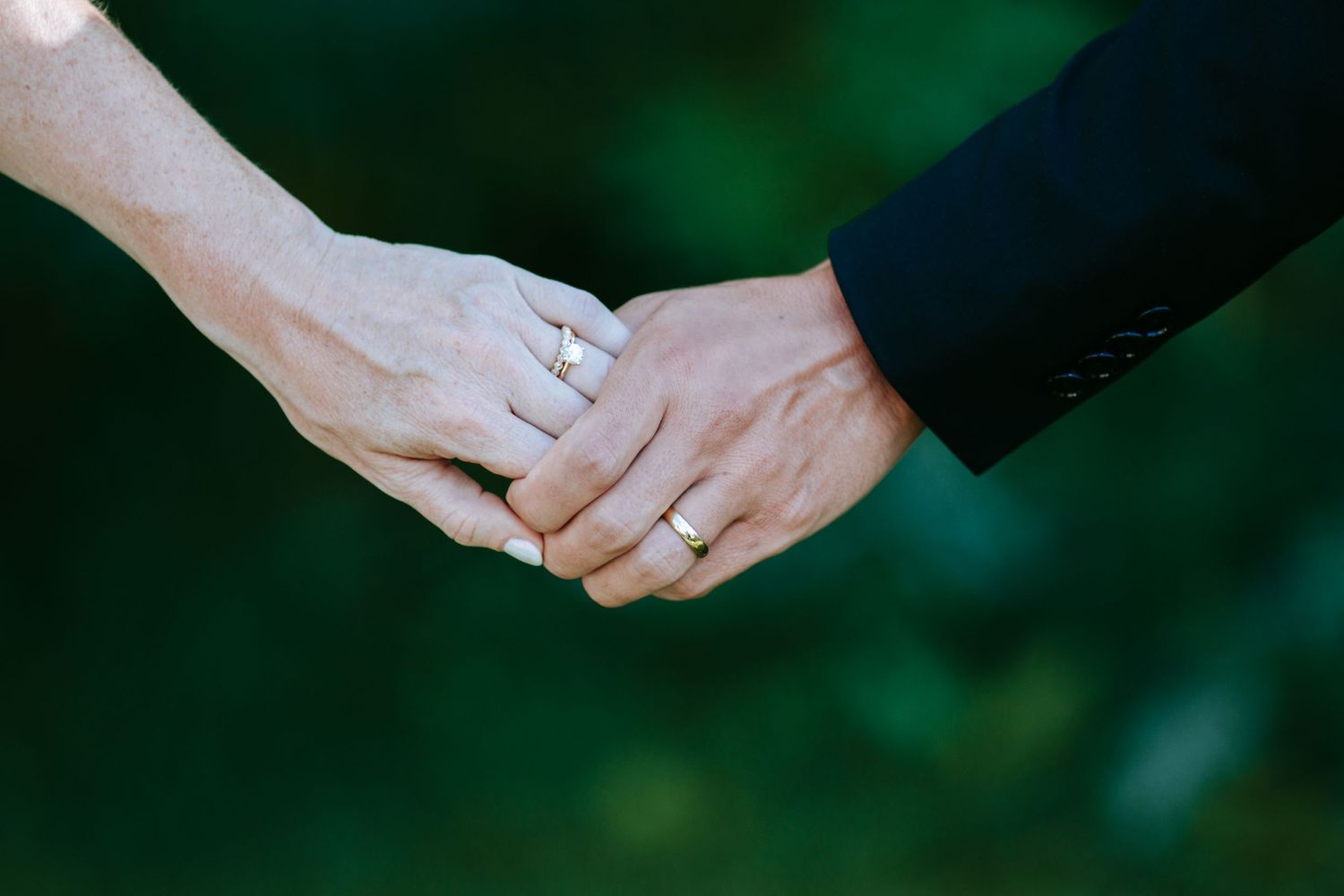 Wedding rings gleam as two hands clasp together against emerald foliage.