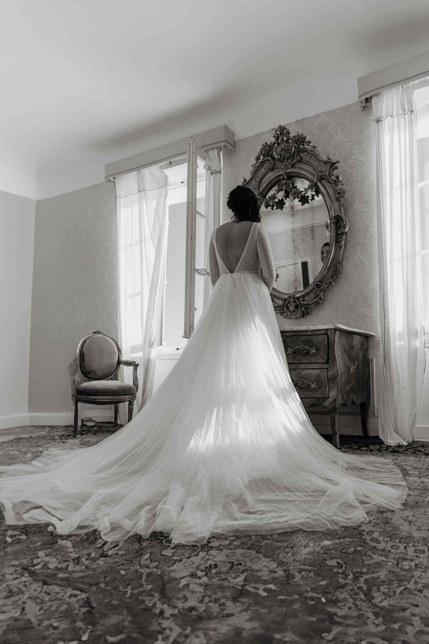 Black and white photograph of an elegant wedding dress with long train viewed from behind in a room with ornate mirror.
