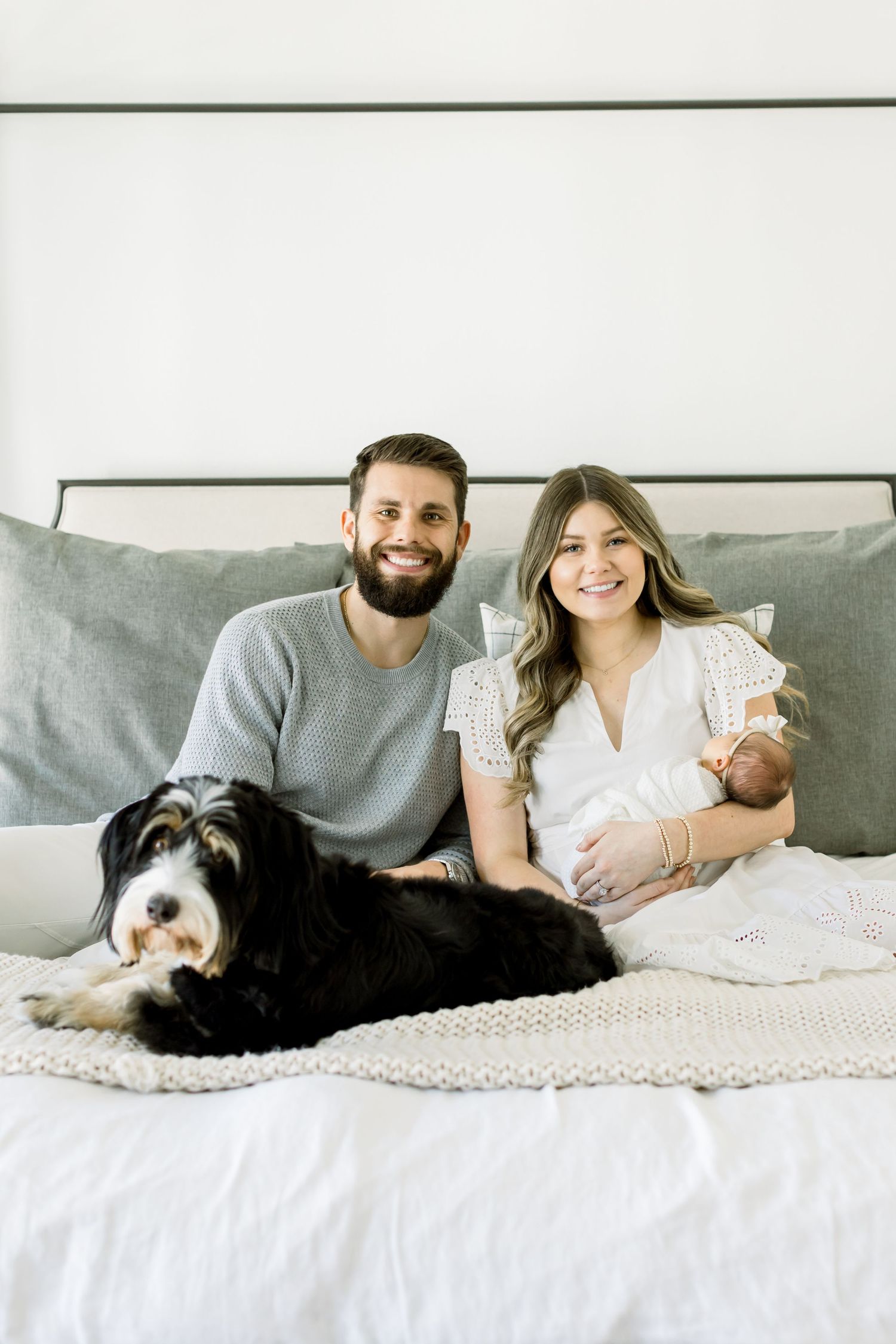 Family portrait with newborn baby and black and white dog on grey couch.