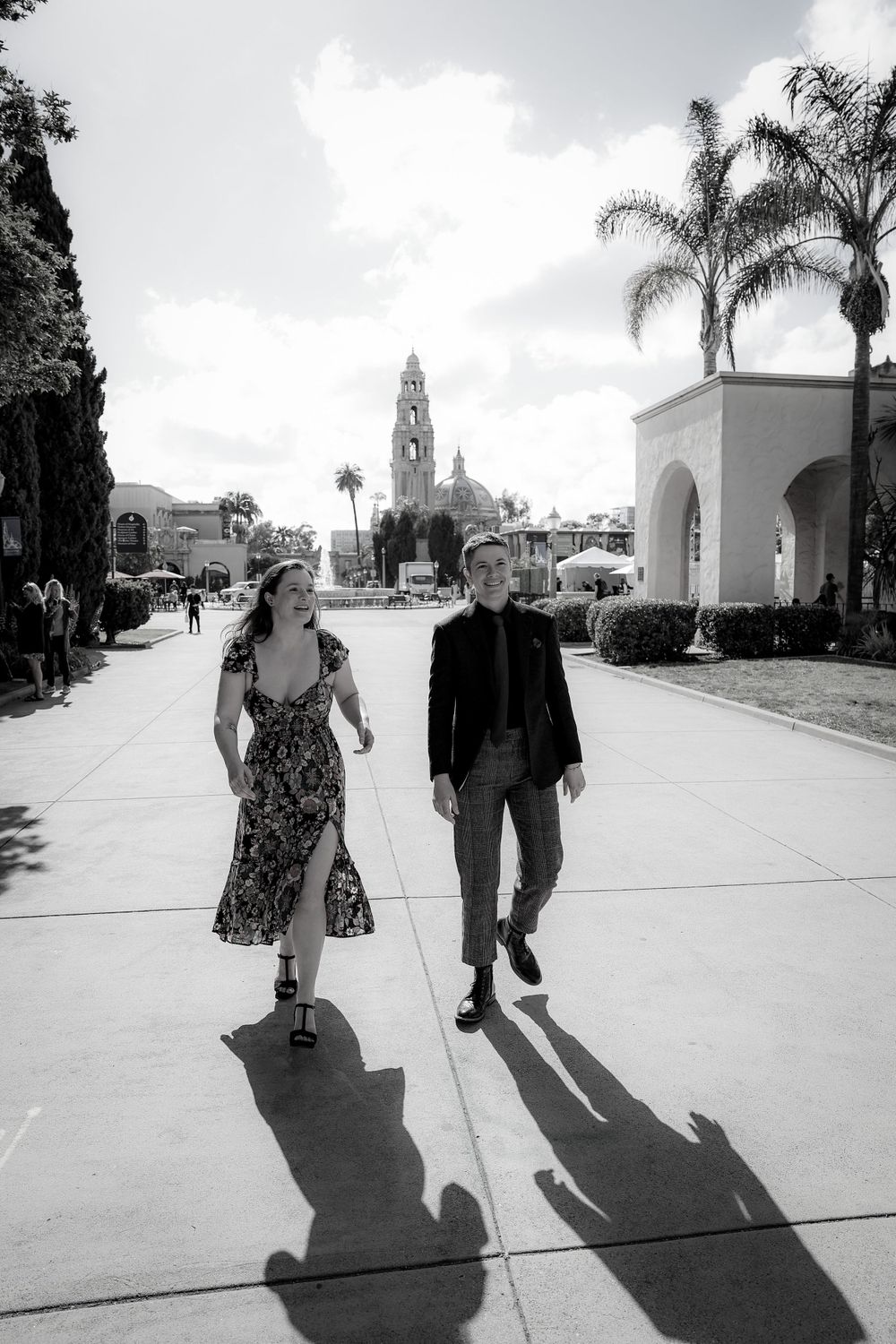 Black and white photo of couple walking on path with Spanish colonial architecture in background.