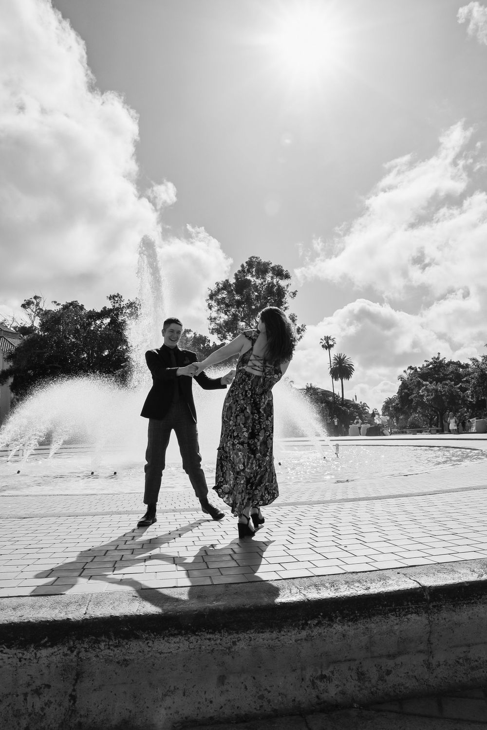 Black and white photo of couple dancing by fountain with dramatic sunlight.