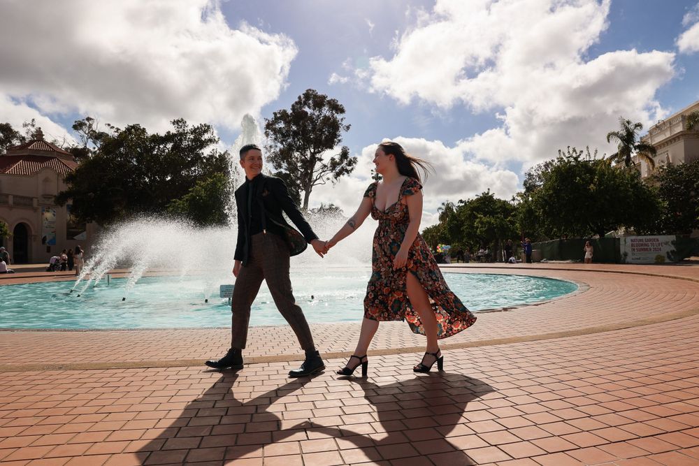 Couple holding hands while dancing near fountain under bright blue sky.
