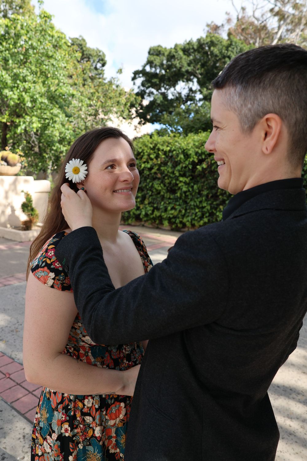 A couple shares a tender moment outdoors with the woman wearing a floral dress while a flower adorns her hair.