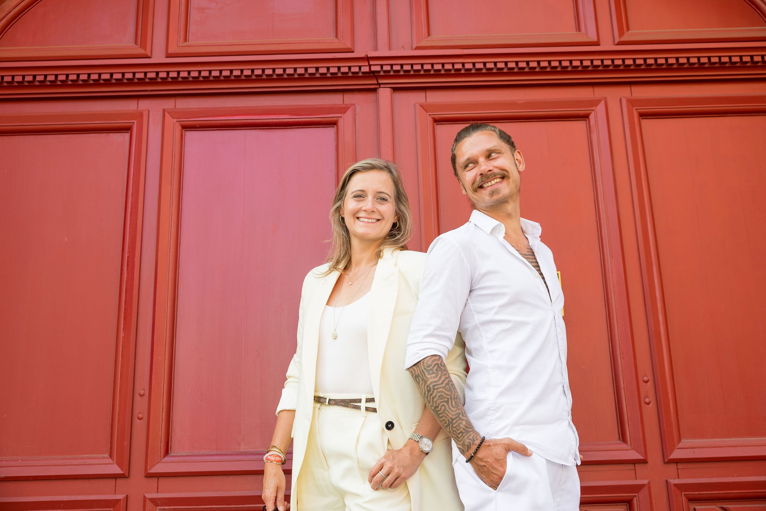 A couple in white summer outfits poses against a bright red wall outdoors. Wedding planner couple from MyCastleWedding in Dresden.
