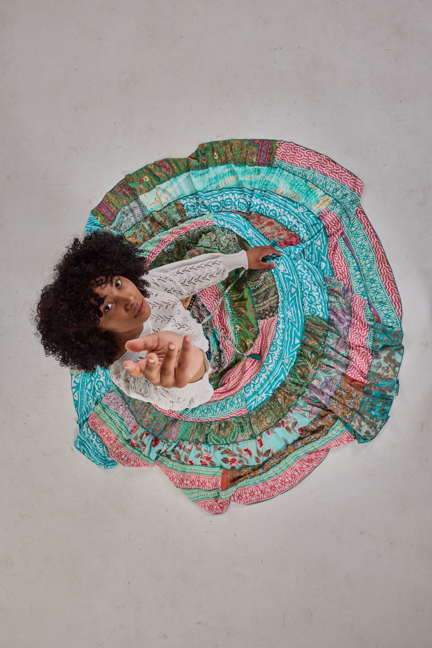 Aerial view of model in white top sitting in colorful patterned maxi skirt against neutral background.