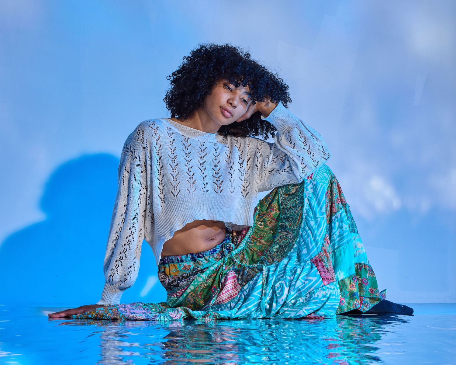 Fashion model in white crochet top and teal patterned skirt poses against blue backdrop in artistic portrait.