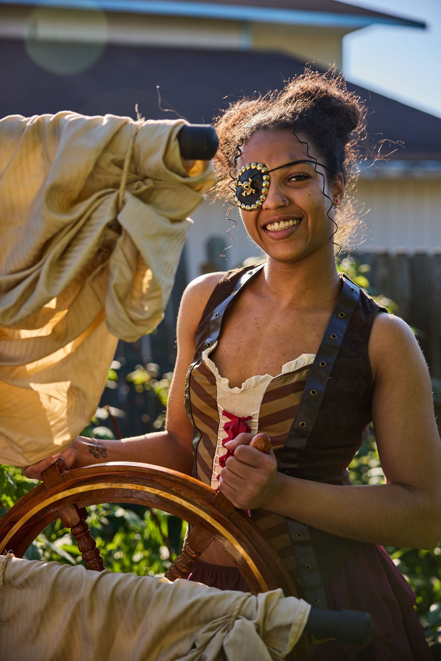 A person wearing ornate steampunk-style goggles and period dress smiles while steering a ships wheel outdoors in sunlight.