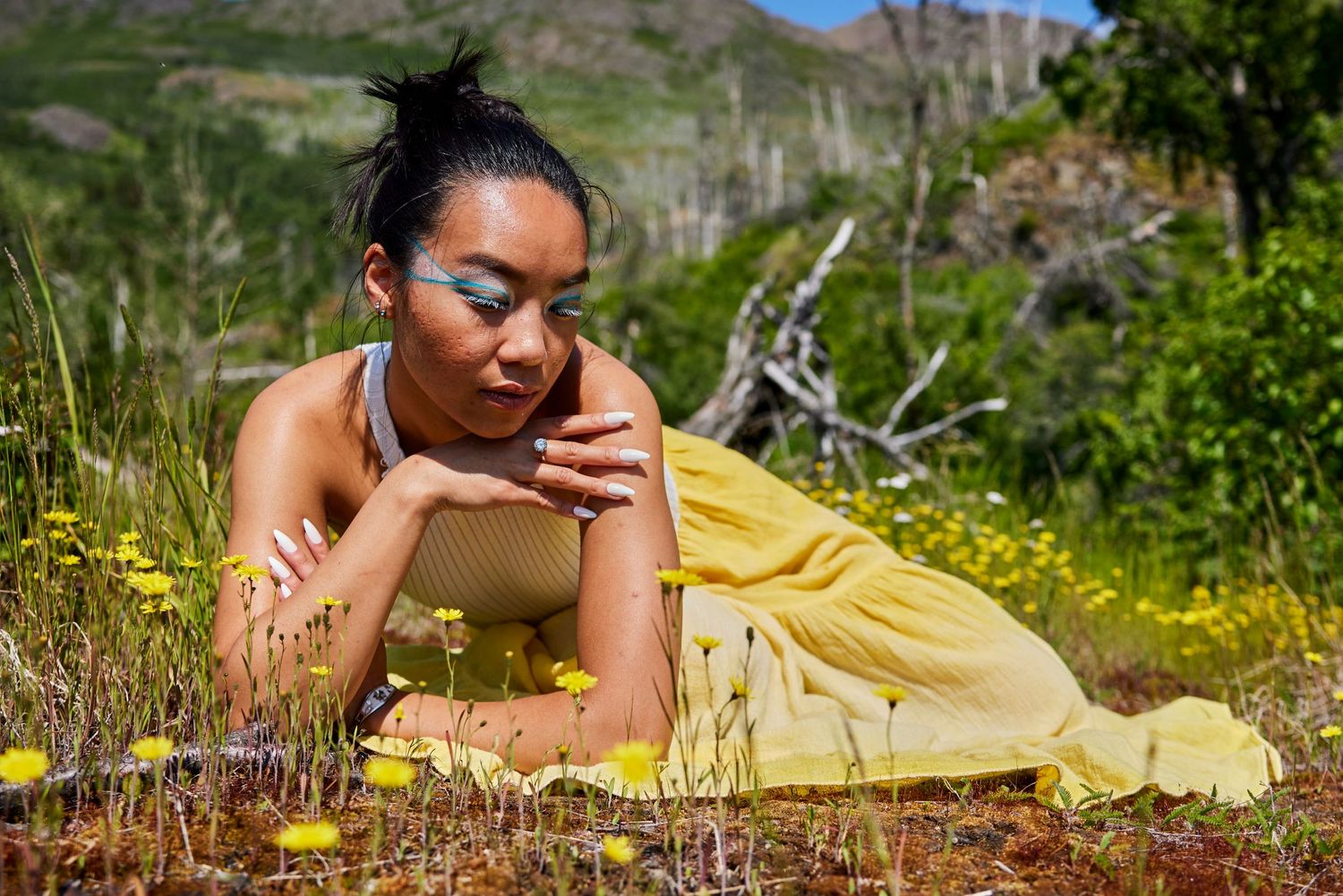 Person in a yellow dress lying on the ground reading outdoors surrounded by wildflowers and natural scenery.