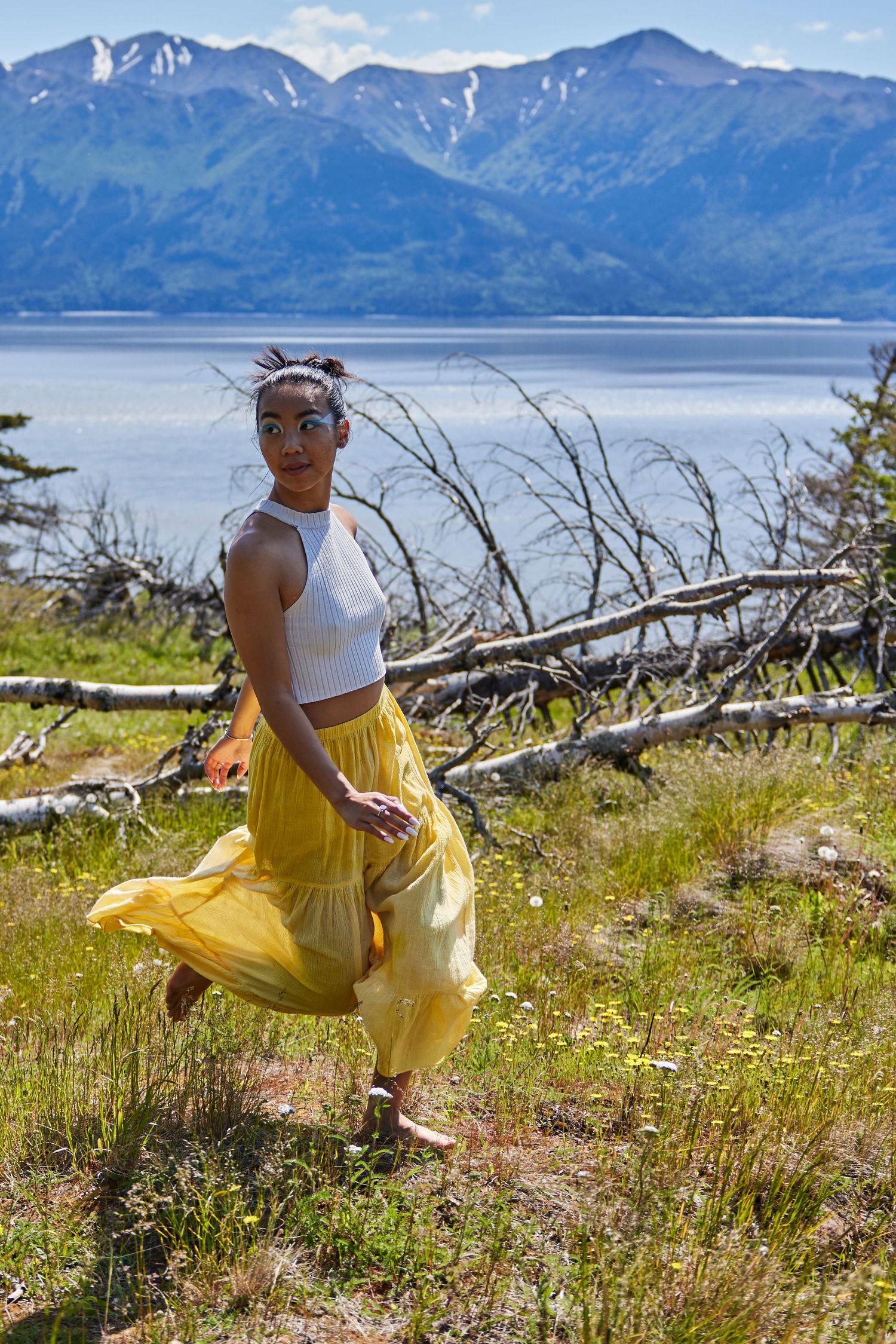 Figure in a yellow flowy skirt dances against a backdrop of mountains and a lake on a sunny day.