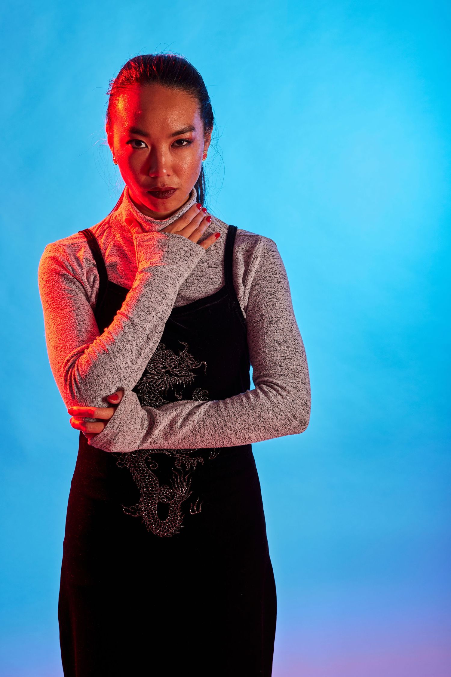 Artistic portrait with red lighting against blue background wearing gray long sleeves and black dress.