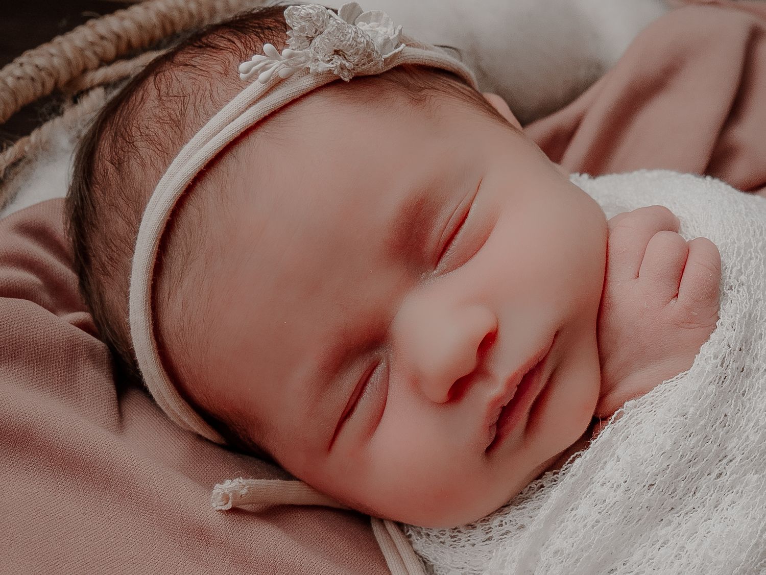 Sleeping newborn wearing lace headband rests on soft pink blanket.