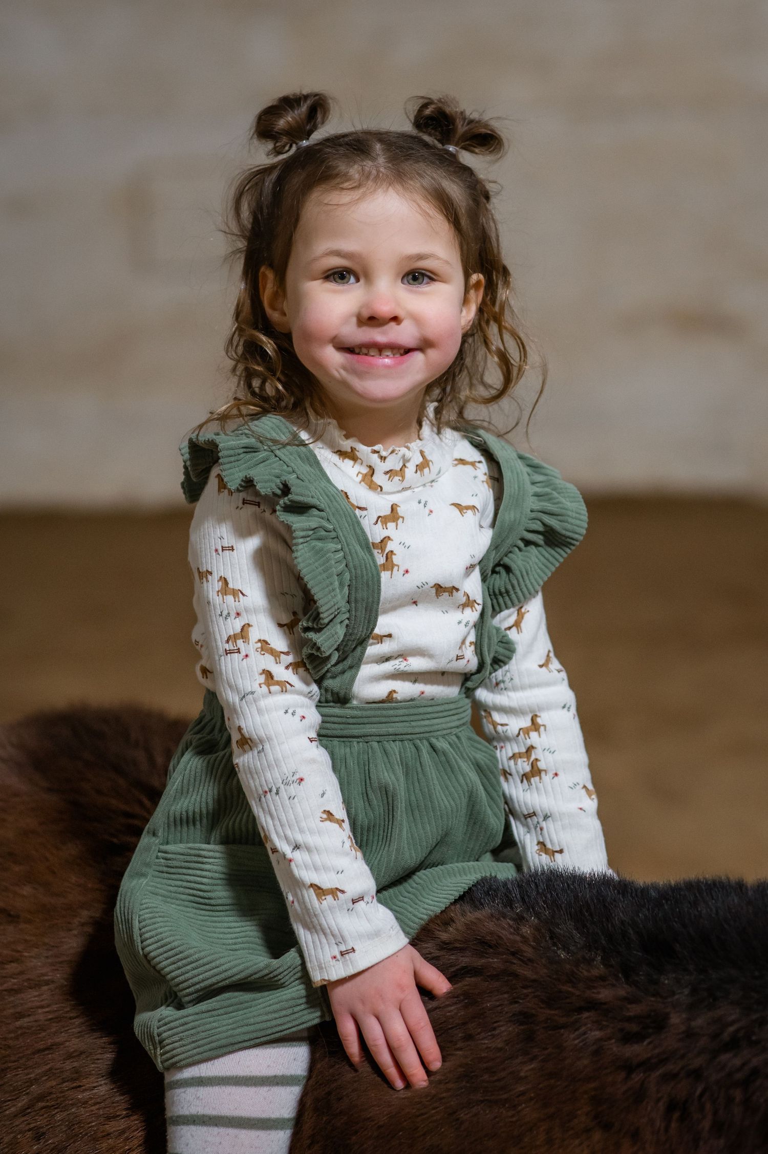 Child in a green corduroy outfit sitting on a furry brown surface with a bright smile.