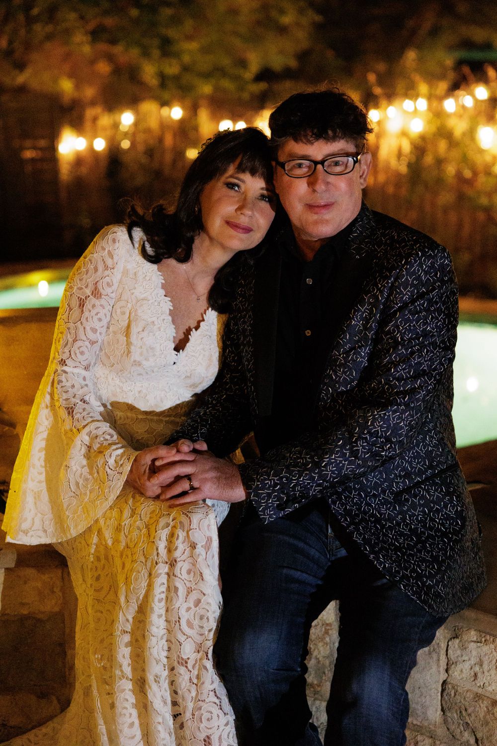 A couple shares an intimate moment by a pool illuminated by garden lights.