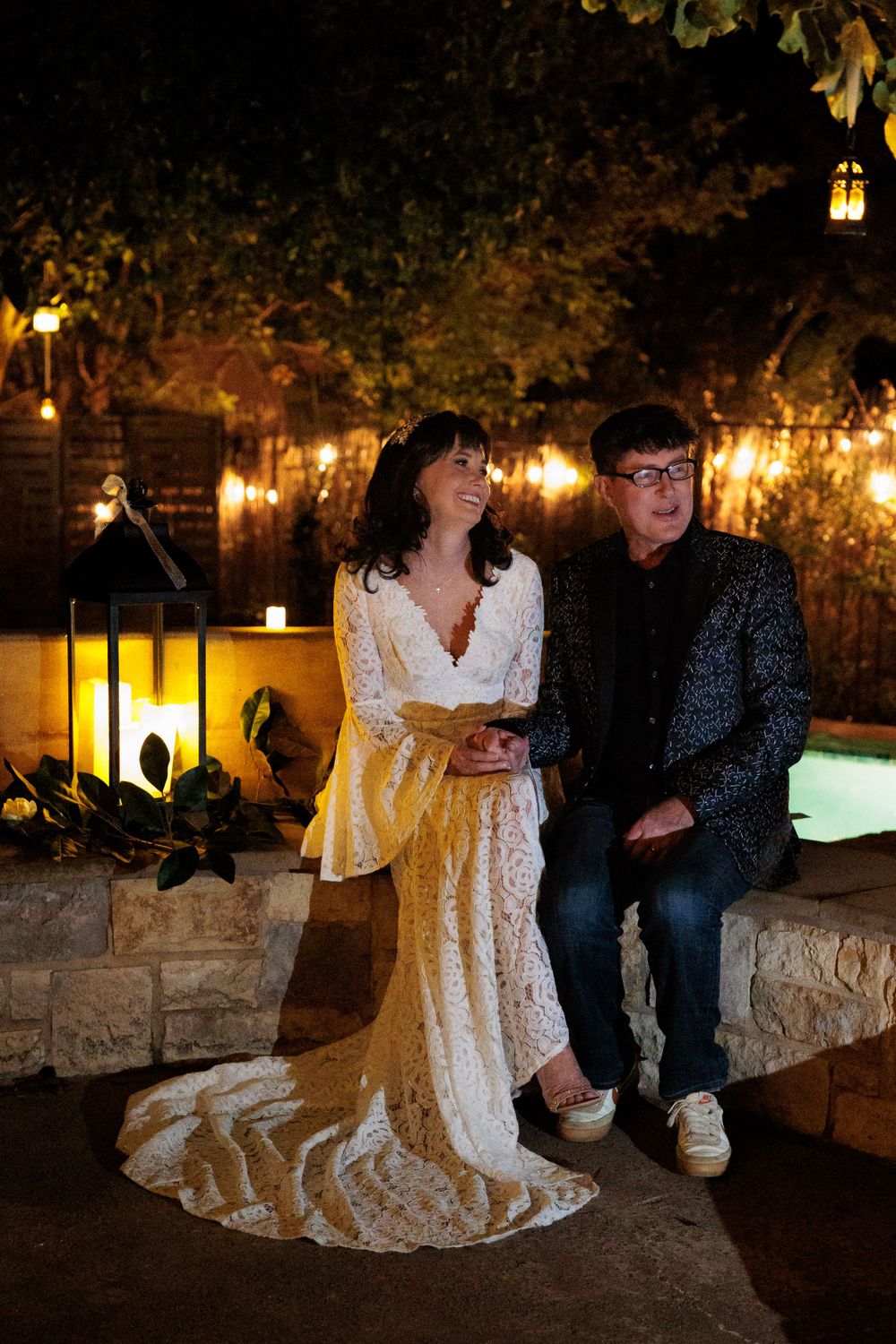 A bride in lace dress and groom in sparkly jacket sit on stone wall under twinkling lights.