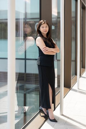 Asian businesswoman in black dress stands confidently with arms crossed beside glass building exterior.