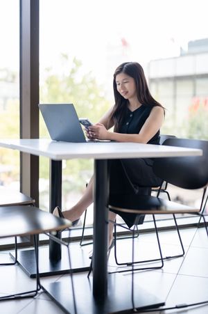 Young Asian businesswoman smiling at smartphone while working on laptop at modern office table near window.