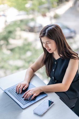 Young businesswoman in black dress typing on laptop at outdoor desk with smartphone nearby.